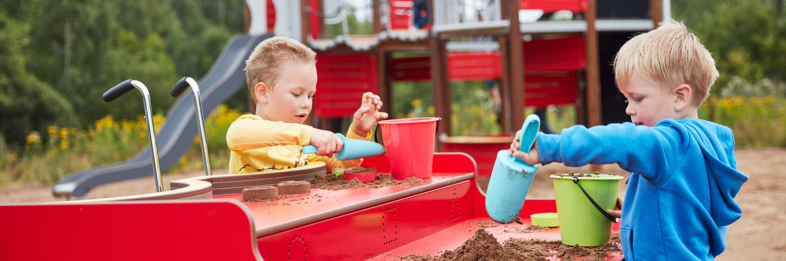 Children play at an outdoor playground sandtable which is elevated. One of the children is a wheelchair user.